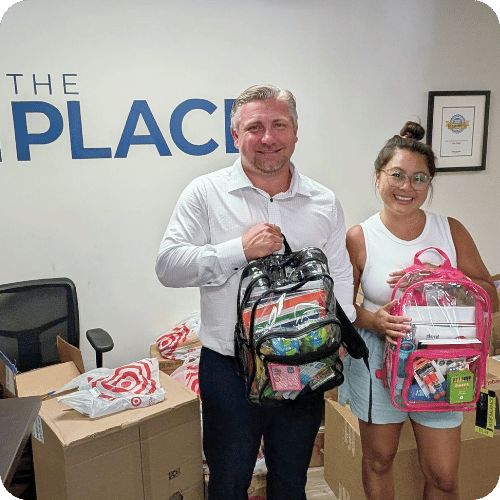 Two people stand indoors holding clear backpacks filled with school supplies. Boxes and bags of supplies are stacked nearby. The wall behind them has part of the word “PLACE” visible.