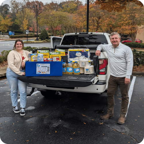 Two people stand by a pickup truck with its tailgate down, displaying boxes and bags of food and supplies in a parking lot on a cloudy day.