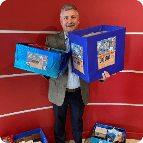 A man in a suit holds two large food donation boxes in front of a red wall, with more boxes on the floor.