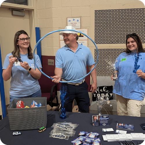 Three adults stand behind a table with snacks, flyers, and toys at an indoor event. One person holds a blue hula hoop. All three wear light blue shirts and smile.