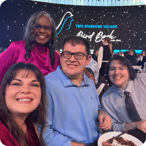 Four people smiling at a banquet table with a “Two Sparrows Village Bird Bash” event sign in the background.