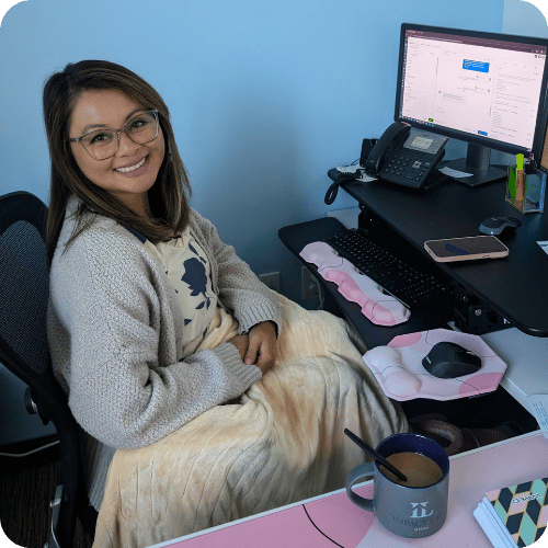 Woman sits at an office desk with a computer, mug, and phone; she is smiling, wearing glasses, and has a blanket over her lap.