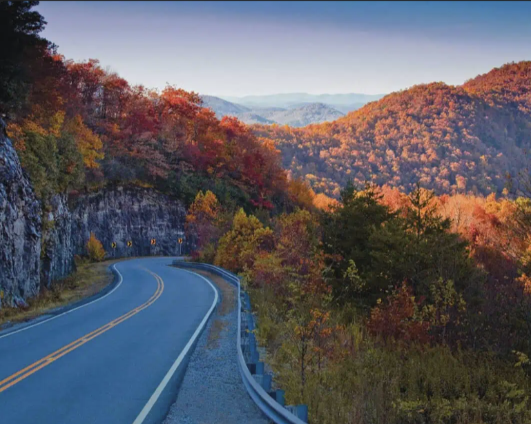 A winding road curves through forested hills with autumn foliage, bordered by a rocky cliff on the left and distant mountains under a clear blue sky.