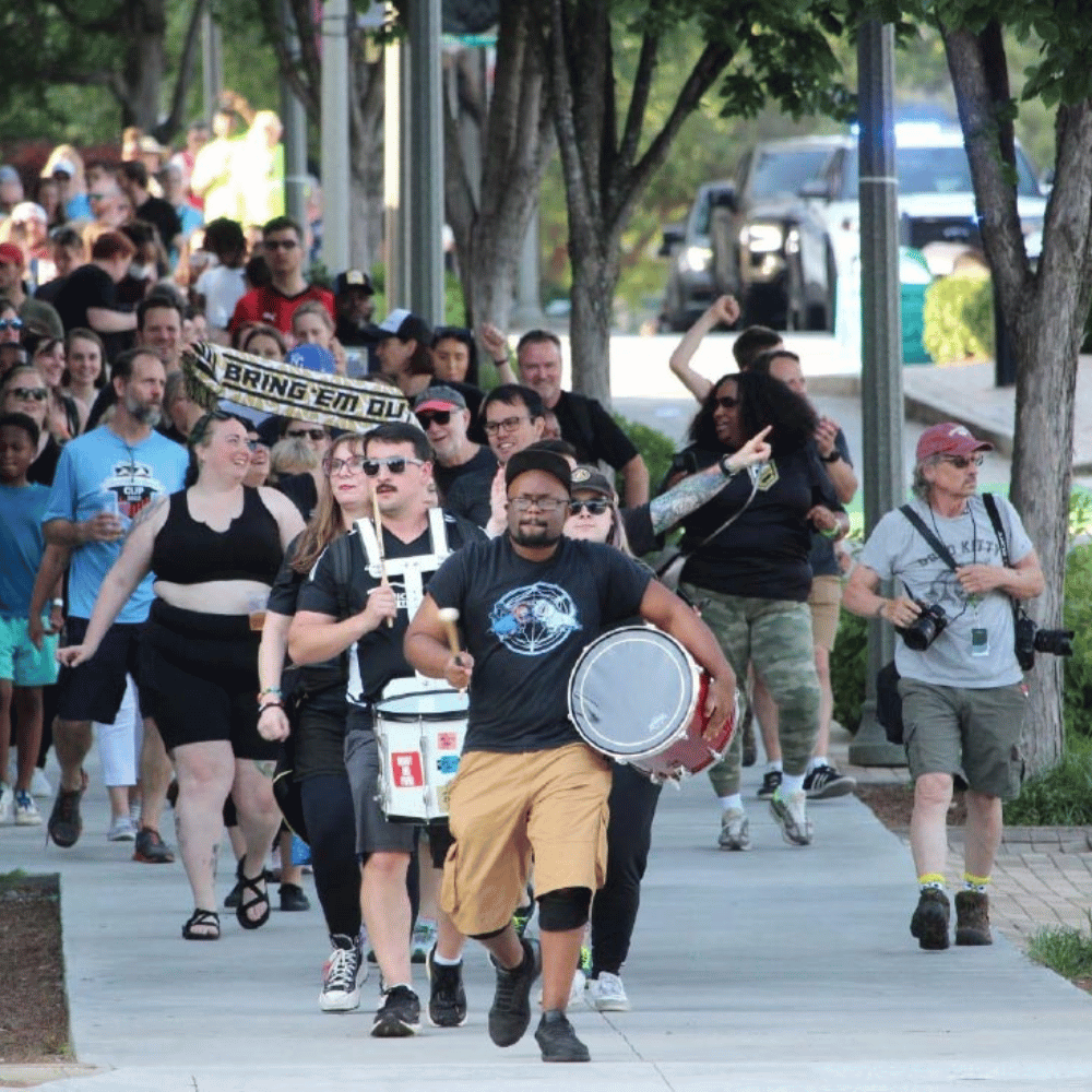 A large group of people marches down a sidewalk; some carry drums and signs, and a police vehicle is visible in the background.