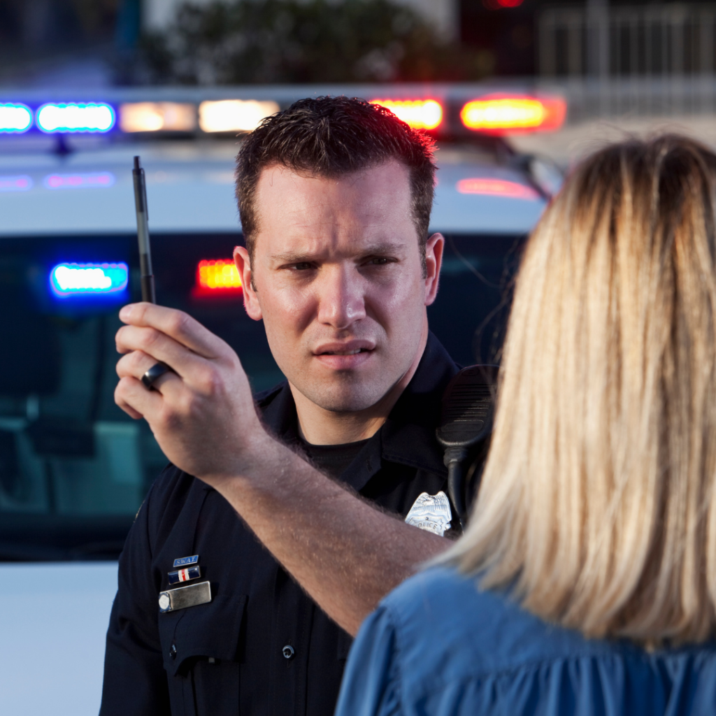 Police officer holds a pen in front of a person during a field sobriety test, with a police car and flashing lights in the background.