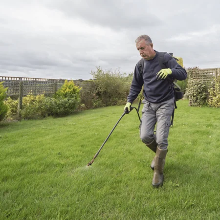 Man wearing gloves and boots sprays lawn with a long-handled applicator in a garden, carrying a backpack sprayer.
