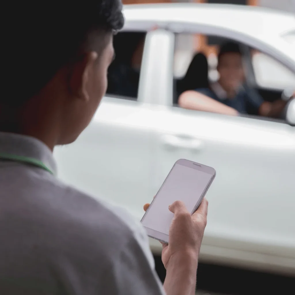 Person holding a smartphone while standing near a white car with people visible inside the car.