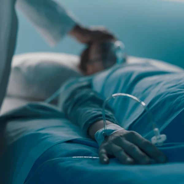 A person lies in a hospital bed with an oxygen mask and IV in their arm, while someone stands nearby touching their head.