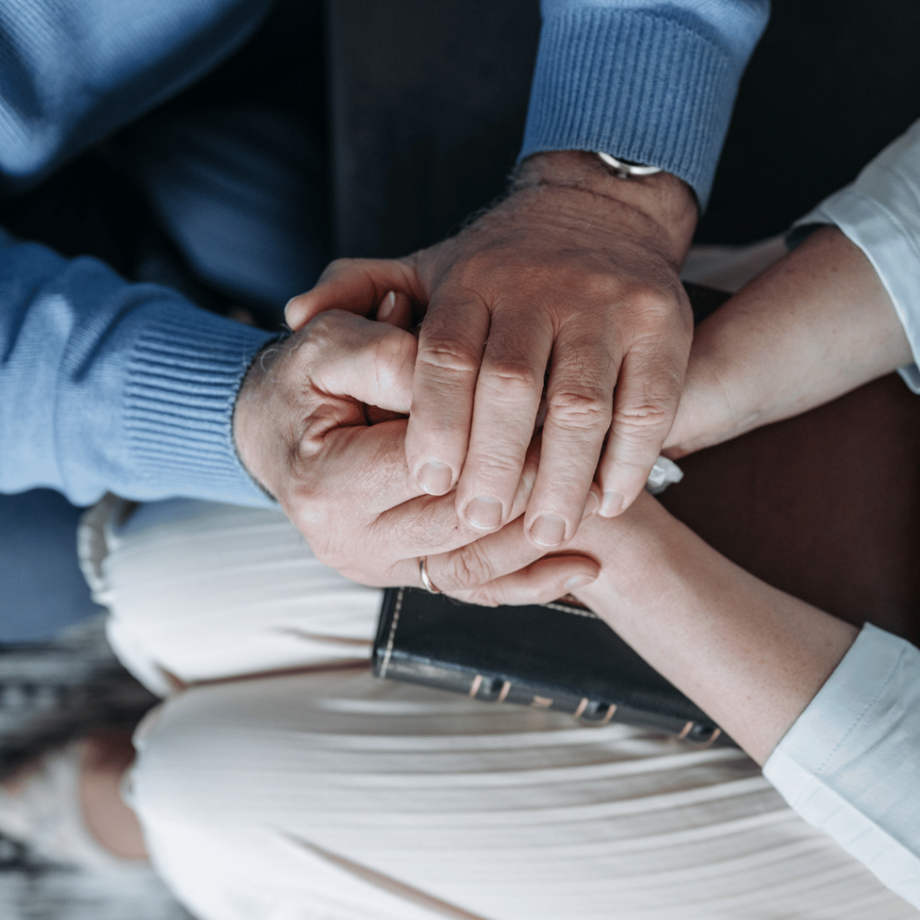 Two people sit close together, one holding a closed book while the other gently holds their hands in a comforting gesture.