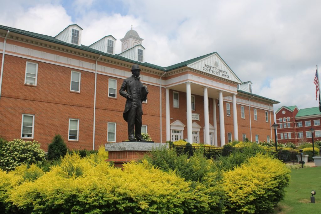 A brick courthouse with white columns stands behind a statue of a man in a garden, with a blue sky and clouds above.