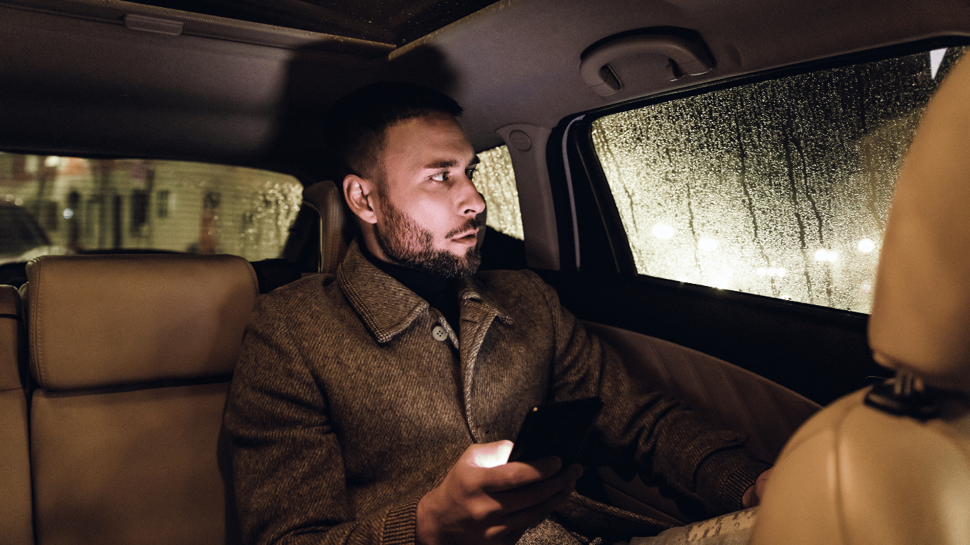 A man sits in the backseat of a car at night, holding a smartphone, looking out the window at rain-soaked glass.