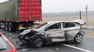 A heavily damaged silver car after a collision with a red truck on a wet highway; debris is scattered on the road.