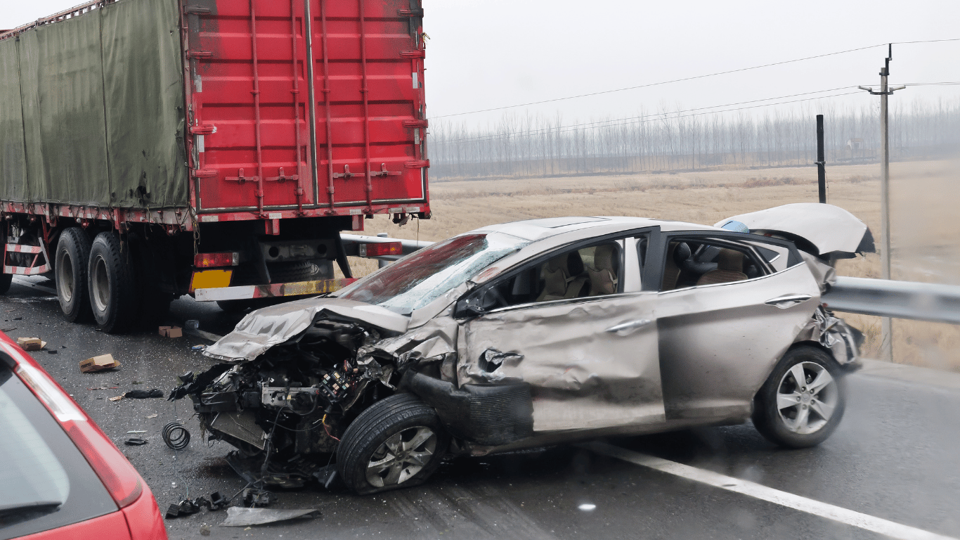 A heavily damaged silver car after a collision with a red truck on a wet highway; debris is scattered on the road.
