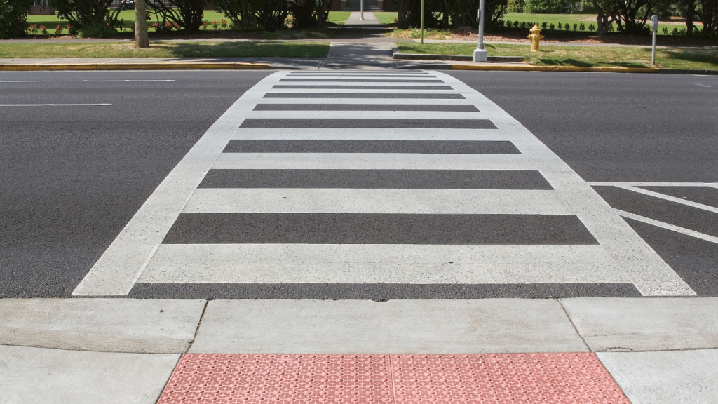 A crosswalk with white stripes spans across an asphalt street. The curb has a textured red ramp for accessibility. Trees and a pathway are visible in the background.