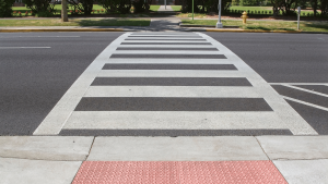 A crosswalk with white stripes spans across an asphalt street. The curb has a textured red ramp for accessibility. Trees and a pathway are visible in the background.