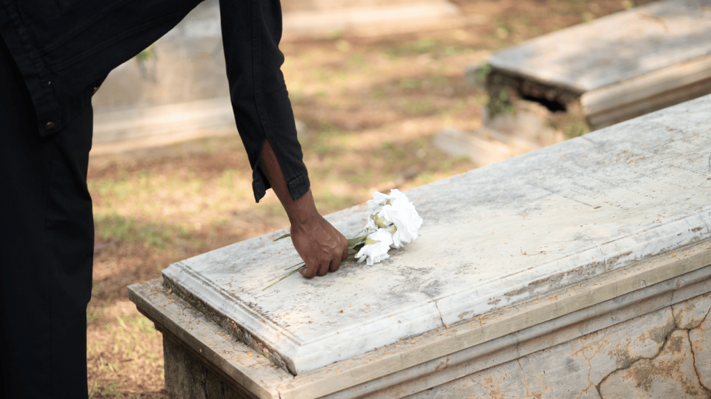 A person places a white bouquet on a stone grave in a cemetery, with sunlight filtering through the trees.