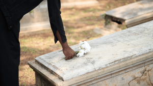 A person places a white bouquet on a stone grave in a cemetery, with sunlight filtering through the trees.