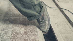 A person wearing dusty work boots steps near a gap in a wooden floor, with scattered debris and a cord visible in the background.
