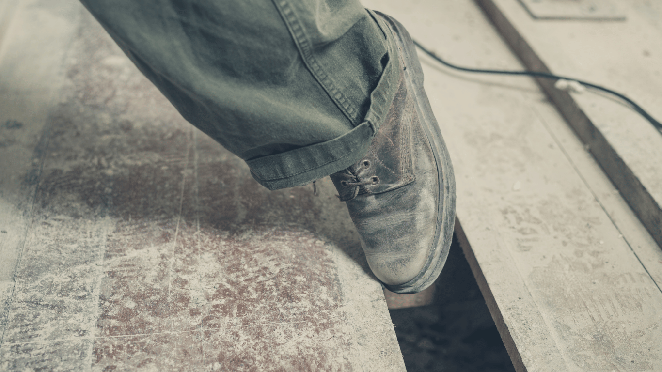 A person wearing dusty work boots steps near a gap in a wooden floor, with scattered debris and a cord visible in the background.