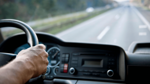 Close-up of a person’s hand on a steering wheel, with a view of a car dashboard and a blurred highway stretching ahead.