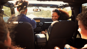 Four people sit inside a car with the roof down, sunlight streaming in, as one woman in the passenger seat smiles at those in the back.
