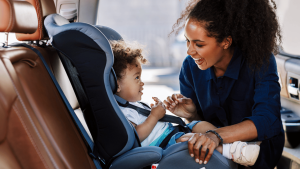 A woman smiles at a toddler sitting in a car seat in the back of a vehicle, checking or adjusting the child’s harness.