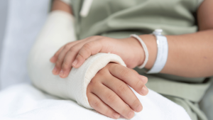 A person wearing a hospital wristband rests their hands on a white surface, with one hand and forearm wrapped in a cast.