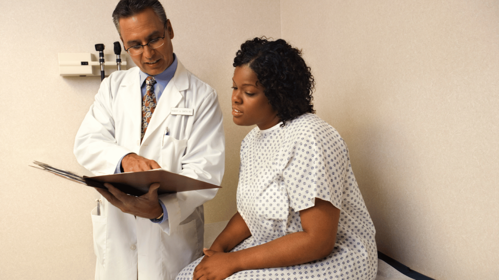A doctor in a white coat explains information from a chart to a patient wearing a hospital gown, seated on an exam table in a medical office.