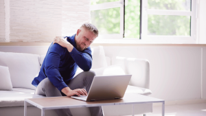 A man in a blue shirt sits on a couch using a laptop, holding his shoulder and appearing to be in pain.