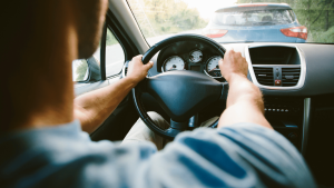 Person driving a car on a sunny day, following another vehicle on a road, with hands on the steering wheel and dashboard gauges visible.