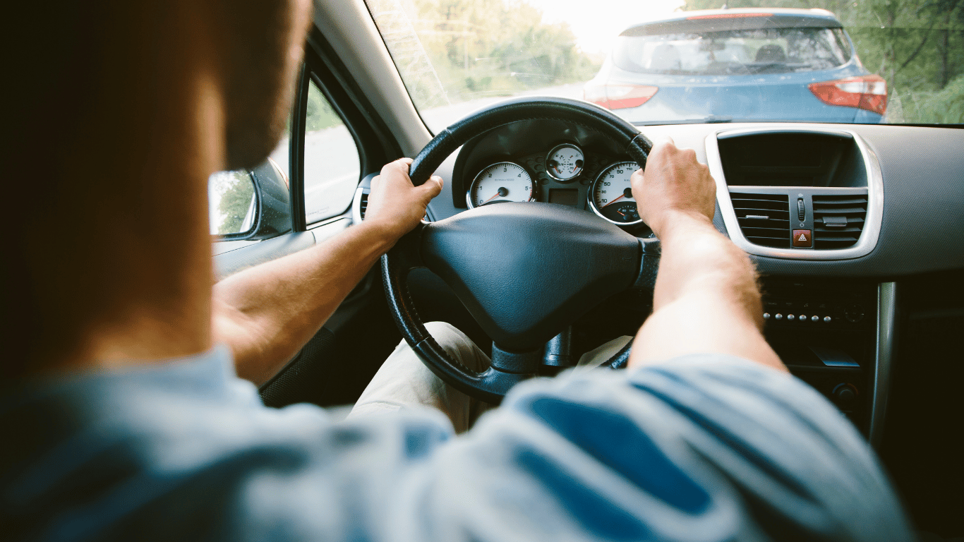 Person driving a car on a sunny day, following another vehicle on a road, with hands on the steering wheel and dashboard gauges visible.