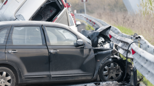 A black car with severe front-end damage is crashed into a metal guardrail; a white vehicle is stacked on top in a multi-car accident.