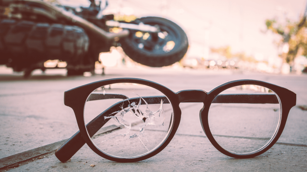 A pair of black-framed glasses with a cracked lens lies on the ground, with a blurred motorcycle lying on its side in the background.