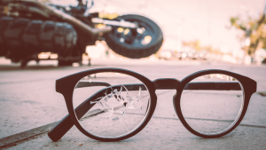 A pair of black-framed glasses with a cracked lens lies on the ground, with a blurred motorcycle lying on its side in the background.
