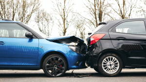 A blue car has collided with the rear of a black car on a road. Both vehicles show visible damage. Trees without leaves are in the background.