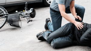 A person lies on the ground beside a fallen bicycle while another person kneels next to them, offering assistance.
