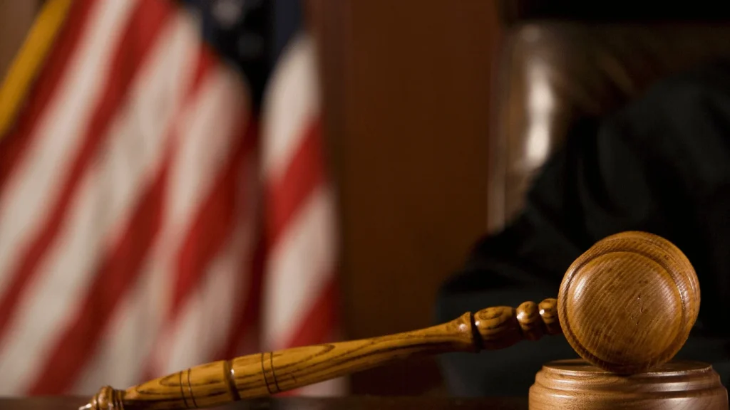 A judge’s gavel rests on a sounding block with part of an American flag and a judge’s robe visible in the background.