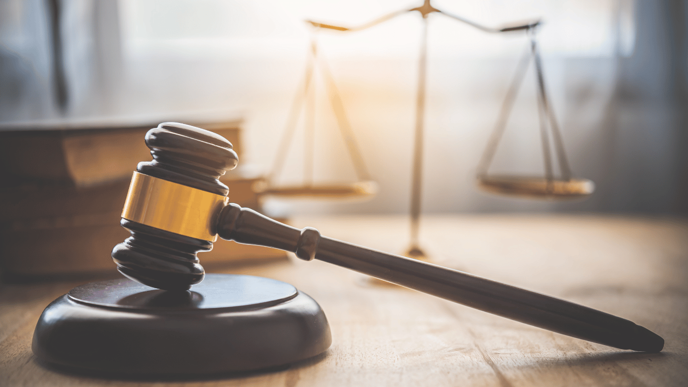 A wooden judge’s gavel rests on a sound block, with a blurred set of balance scales in the background on a wooden table.