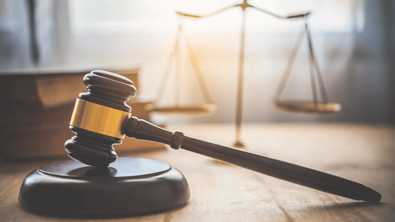 A wooden judge’s gavel rests on a sound block, with a blurred set of balance scales in the background on a wooden table.
