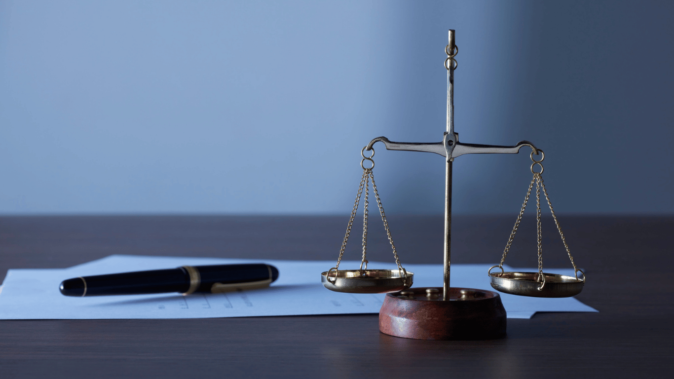 A brass balance scale sits on a wooden desk next to a black pen and a sheet of paper against a plain blue background.