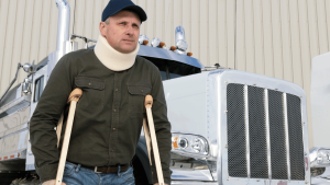 Man with a neck brace and crutches stands in front of a large white semi truck, wearing a dark shirt and blue cap.