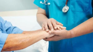 A healthcare worker in blue scrubs gently holds the hand of a patient, offering support and comfort in a clinical setting.
