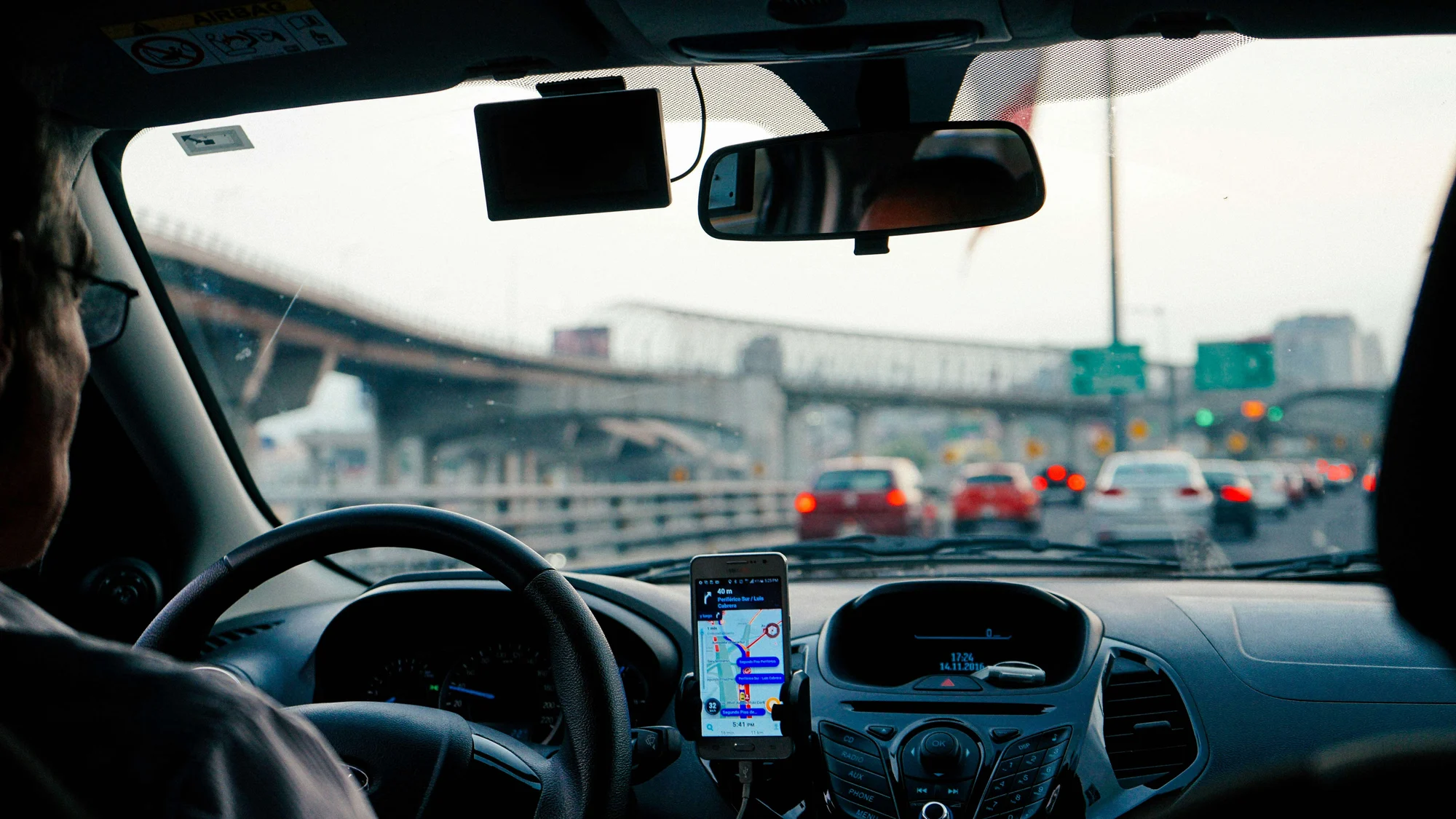 A person drives a car in heavy traffic with a GPS navigation app displayed on a smartphone mounted to the dashboard.