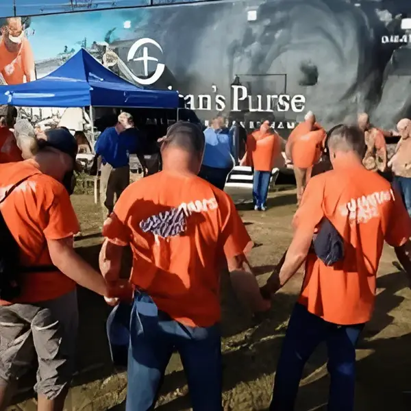 A group of people wearing orange shirts stand in a circle holding hands outdoors, with a Samaritan’s Purse truck and blue tent visible in the background.