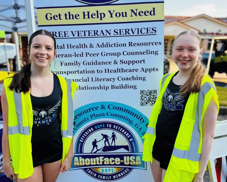 Two smiling girls wearing yellow safety vests stand in front of a sign for free veteran services at an outdoor event.