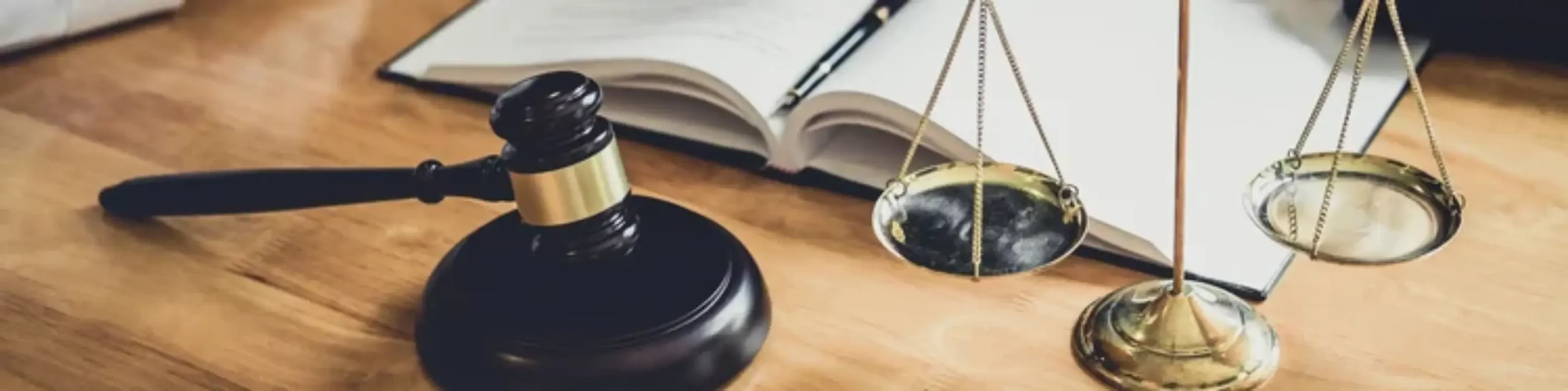 A wooden gavel, brass balance scale, and open notebook rest on a wooden desk, symbolizing law and justice.