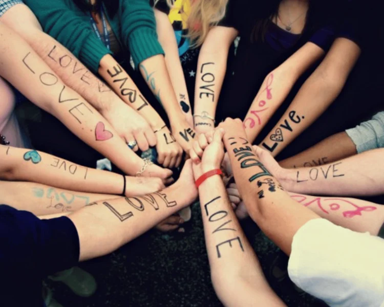 A group of people stand in a circle with their arms extended, each arm featuring the word “LOVE” written on the skin in marker.