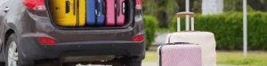 A car with its trunk open shows several colorful suitcases inside. Two more suitcases stand on the ground behind the car, with greenery visible in the background.