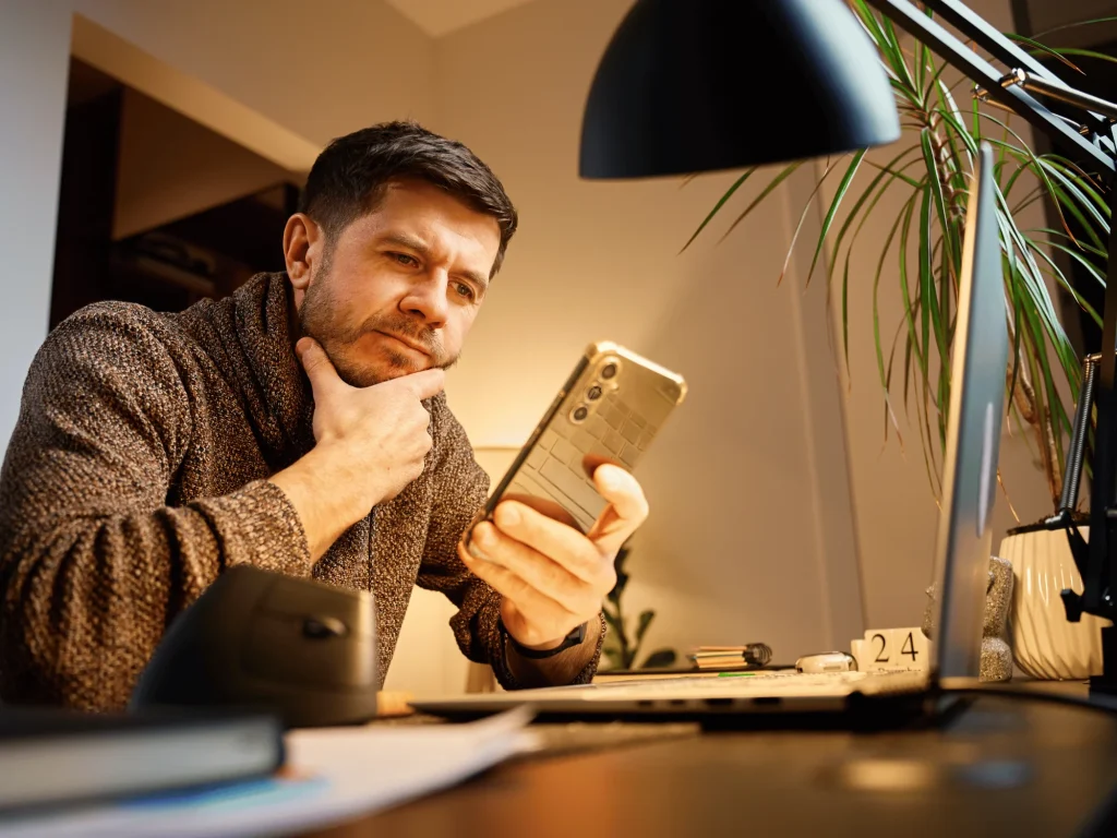 Man sits at a desk, looking thoughtfully at his phone, with an open laptop, lamp, and plant in the background.
