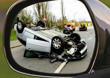 A flipped car with severe damage is seen through a side mirror at an accident scene, with emergency vehicles and responders in the background.
